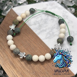 Puede incluir: Un brazalete de cuentas verdes y blancas con dijes de tortuga plateados. El brazalete está atado con un cordón verde y tiene un nudo en el extremo. El brazalete está sobre una superficie de madera con un logotipo de unicornio que dice "unicorn - beading company - ".