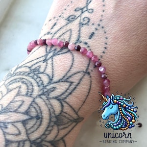 Op de afbeelding: Een roze en paarse edelsteen armband met een zilveren sluiting. De armband zit om de pols van een persoon met een tatoeage.