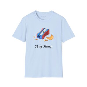 May include: A light blue short-sleeve t-shirt featuring a graphic of a red and blue pencil sharpener with orange pencil shavings. The text "Stay Sharp" is printed in black below the graphic. The shirt has a crew neckline and a standard fit.