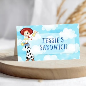 Op de afbeelding: Een gevouwen kaart met het personage Jessie uit Toy Story, tegen een blauwe lucht en wolkenachtergrond. De tekst "Jessie's Sandwich" staat in een witte rechthoek. De kaart ligt op een houten oppervlak.