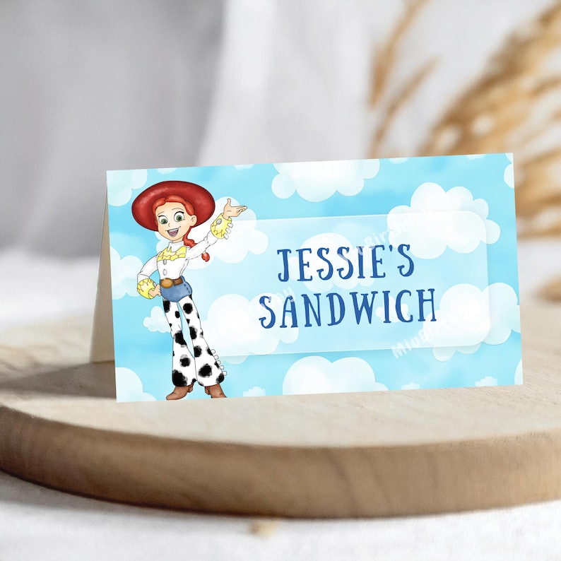 Op de afbeelding: Een gevouwen kaart met het personage Jessie uit Toy Story, tegen een blauwe lucht en wolkenachtergrond. De tekst "Jessie's Sandwich" staat in een witte rechthoek. De kaart ligt op een houten oppervlak.