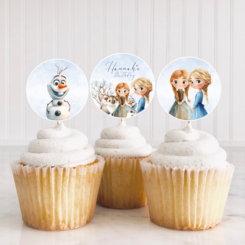 Puede incluir: Tres cupcakes con glaseado blanco y adornos decorativos con personajes de una popular pel&iacute;cula de animaci&oacute;n. Un adorno muestra un mu&ntilde;eco de nieve, otro tiene el texto "Hannah's Birthday", y el tercero muestra a dos chicas con vestidos azules.