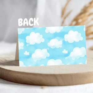 Op de afbeelding: Een gevouwen kaart met een blauwe lucht en wit wolkenpatroon op de achterkant. Het woord "BACK" staat in het wit bovenaan. De kaart wordt gepresenteerd op een houten oppervlak.