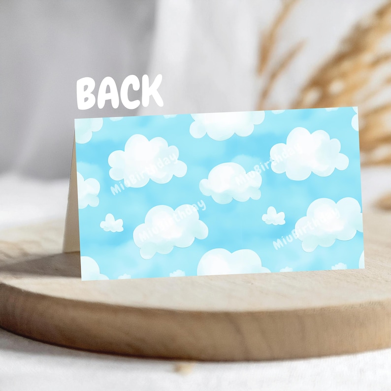 Op de afbeelding: Een gevouwen kaart met een blauwe lucht en wit wolkenpatroon op de achterkant. Het woord "BACK" staat in het wit bovenaan. De kaart wordt gepresenteerd op een houten oppervlak.