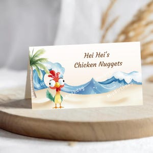 Op de afbeelding: Gevouwen kaart met de tekst "Hei Hei's Chicken Nuggets" boven een cartoonhaan op een strand met golven en een palmboom. De kaart ligt op een houten oppervlak.