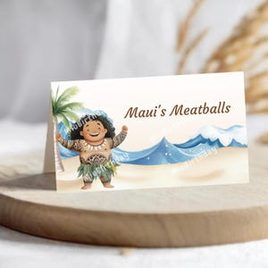 Op de afbeelding: Een gevouwen kaart met een cartoon illustratie van Maui uit de film Moana. De kaart heeft de tekst "Maui's Meatballs" en toont een strandsc&egrave;ne met een palmboom en golven. De kaart ligt op een houten oppervlak.