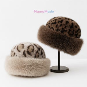 May include: Two leopard print hats with fur trim. One hat is displayed on a black stand. The hats have a round crown and a wide fur band around the base. The text "MamaMade" is in the upper left corner.