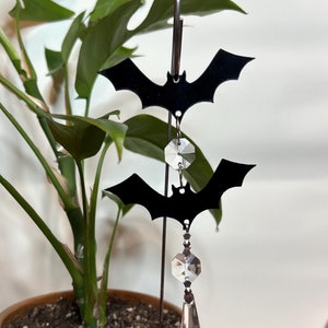 May include: Two black bat cutouts hang from a metal rod with crystal prism accents. The bats are suspended from the rod with a clear crystal prism hanging below each bat.