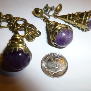 May include: A pair of gold-tone clip-on earrings with purple amethyst stones. The earrings feature a cone-shaped design with a wire-wrapped detail.