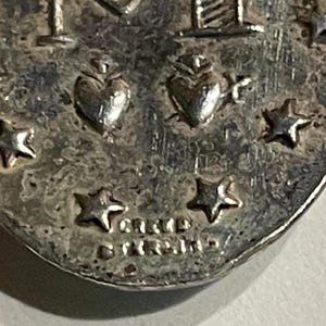 May include: A silver-toned oval pendant featuring an embossed letter M, two hearts, and multiple stars. The surface has a textured, aged appearance. The text "CREED" and "STERLING" is clearly stamped at the bottom of the metal surface.