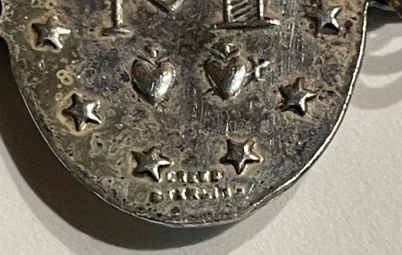 May include: A silver-toned oval pendant featuring an embossed letter M, two hearts, and multiple stars. The surface has a textured, aged appearance. The text "CREED" and "STERLING" is clearly stamped at the bottom of the metal surface.