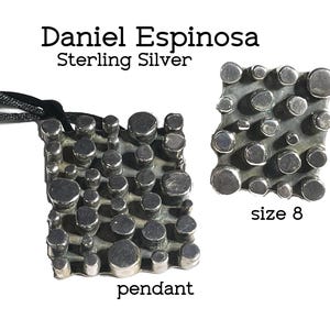 May include: A Daniel Espinosa sterling silver pendant and a size 8 ring. Both pieces feature a dark, oxidized metal base with numerous raised, polished silver circular studs of varying sizes across the rectangular surfaces. A black cord is attached.