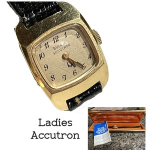 May include: A gold-toned Bulova Accutron watch with a square face and black leather band. The watch face has black Roman numeral hour markers and a white background. The watch is in a box with a white and blue label that says "To the Bulova Accutron Dealer".