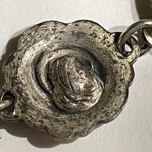 May include: A silver-toned metal link from a rosary featuring a circular, scalloped-edge medallion with a raised relief depiction of a figure in a hooded cloak. The metal surface has a textured, aged finish and connects to other chain links.