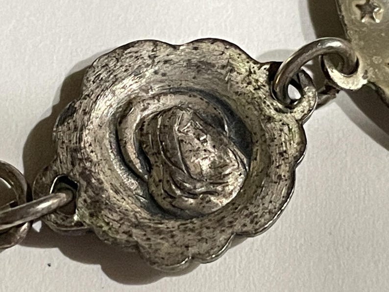 May include: A silver-toned metal link from a rosary featuring a circular, scalloped-edge medallion with a raised relief depiction of a figure in a hooded cloak. The metal surface has a textured, aged finish and connects to other chain links.