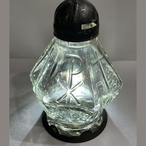 May include: A clear glass memorial candle holder with a faceted, diamond-like shape. The glass features embossed religious symbols, including a Chi-Rho and a cross. It is topped with a black metal dome lid and sits on a black circular base.