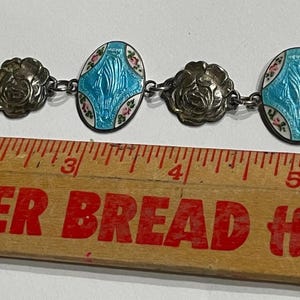 May include: A silver-toned link bracelet featuring three oval charms with blue enamel and floral accents alternating with three embossed rose-shaped links. The bracelet is positioned above a wooden ruler, showing a total length of approximately 16.5 cm.