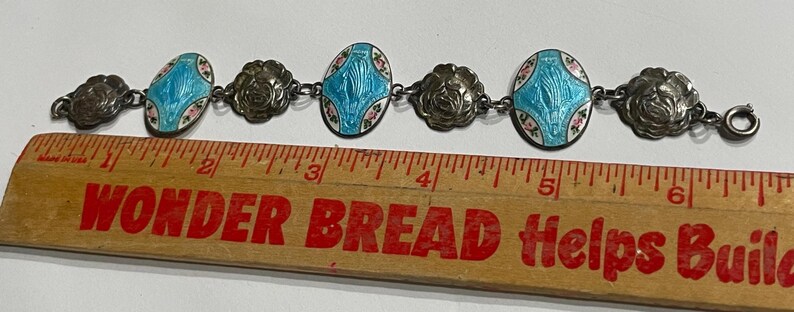 May include: A silver-toned link bracelet featuring three oval charms with blue enamel and floral accents alternating with three embossed rose-shaped links. The bracelet is positioned above a wooden ruler, showing a total length of approximately 16.5 cm.