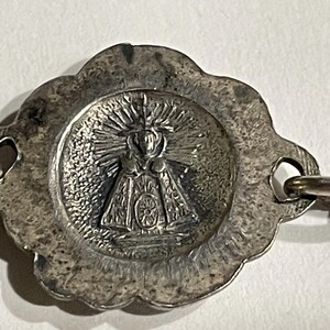 May include: A silver-toned metal charm featuring a central circular medallion with a relief image of a figure in a robe surrounded by radiating sunburst lines. The medallion has a scalloped edge and two side loops for attachment to a chain.