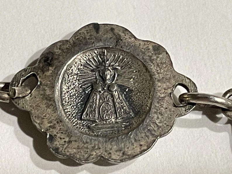 May include: A silver-toned metal charm featuring a central circular medallion with a relief image of a figure in a robe surrounded by radiating sunburst lines. The medallion has a scalloped edge and two side loops for attachment to a chain.