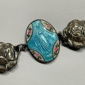 May include: A silver-toned bracelet featuring a central oval charm with a light blue enamelled figure of the Virgin Mary, accented by small painted pink roses on a white border, connected to sculpted silver-toned rose-shaped metal links.