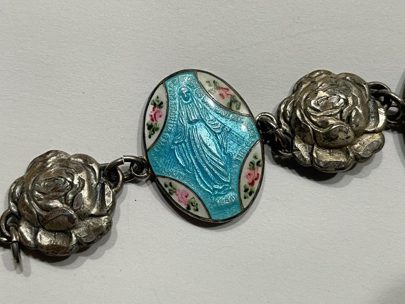 May include: A silver-toned bracelet featuring a central oval charm with a light blue enamelled figure of the Virgin Mary, accented by small painted pink roses on a white border, connected to sculpted silver-toned rose-shaped metal links.
