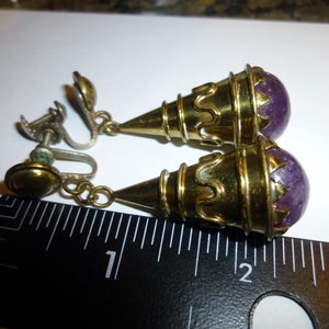 May include: A pair of gold tone clip-on earrings with purple stone accents. The earrings are approximately 2.5 cm long.