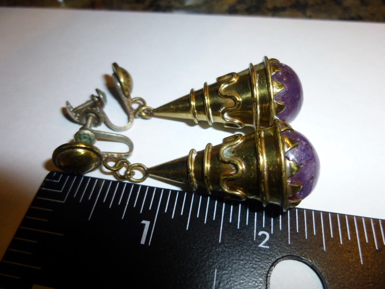 May include: A pair of gold tone clip-on earrings with purple stone accents. The earrings are approximately 2.5 cm long.