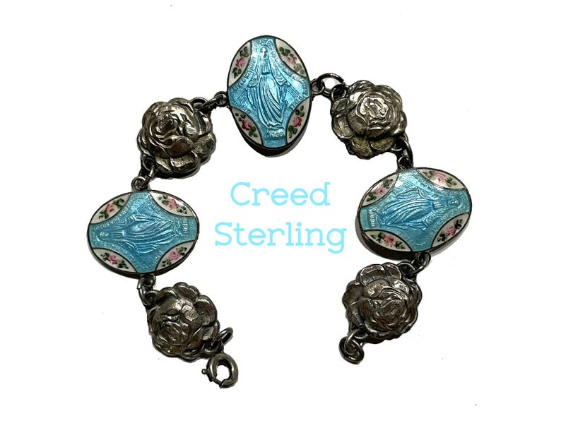 May include: A sterling silver bracelet featuring three oval-shaped charms with light blue enamel and a central figure, alternating with four silver rose-shaped links. The text Creed Sterling is printed in light blue in the centre of the image.