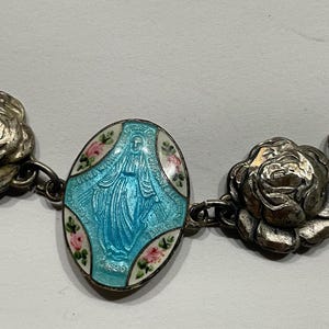 May include: A silver-toned bracelet featuring a central oval charm with a light blue enamel depiction of the Virgin Mary, accented by small painted pink roses, flanked by two detailed silver-toned rose-shaped metal links on each side.