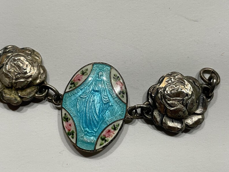 May include: A silver-toned bracelet featuring a central oval charm with a light blue enamel depiction of the Virgin Mary, accented by small painted pink roses, flanked by two detailed silver-toned rose-shaped metal links on each side.