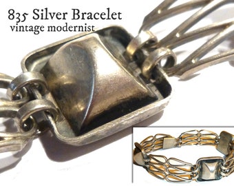 May include: A silver bracelet with a modernist design. The bracelet is made of interlocking links and has a square clasp with a pyramid-shaped top.