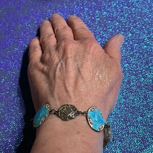 May include: A silver-toned link bracelet featuring alternating oval charms with bright blue enamel and floral accents, interspersed with a central circular metal flower charm. The bracelet is constructed with a series of connected metal links.
