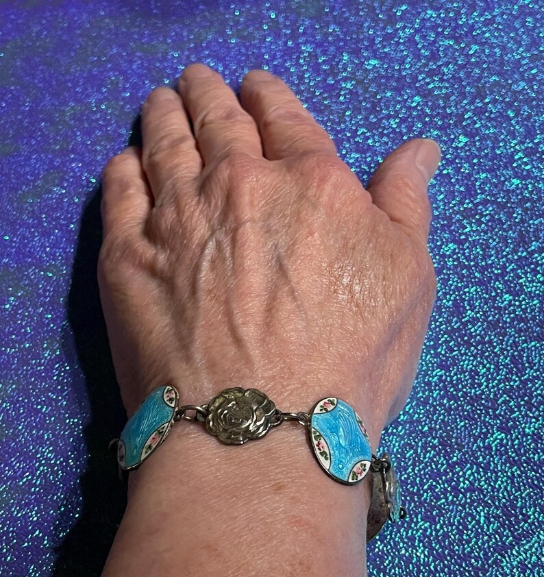 May include: A silver-toned link bracelet featuring alternating oval charms with bright blue enamel and floral accents, interspersed with a central circular metal flower charm. The bracelet is constructed with a series of connected metal links.
