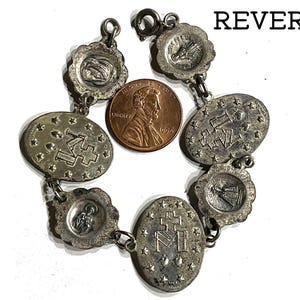 May include: A silver-toned metal charm bracelet featuring multiple oval and scalloped religious medallions with embossed symbols and figures, including the letter M and a cross, displayed next to a copper-coloured 1994 United States penny.