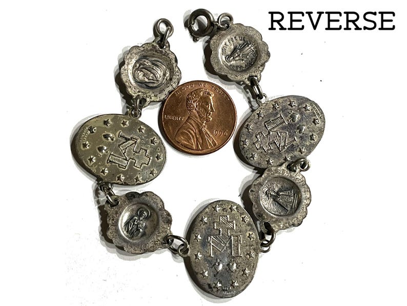 May include: A silver-toned metal charm bracelet featuring multiple oval and scalloped religious medallions with embossed symbols and figures, including the letter M and a cross, displayed next to a copper-coloured 1994 United States penny.