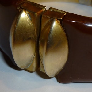 May include: A brown plastic bangle bracelet with two gold-toned metal accents. The accents are shaped like leaves and are attached to the bracelet with a hinge.