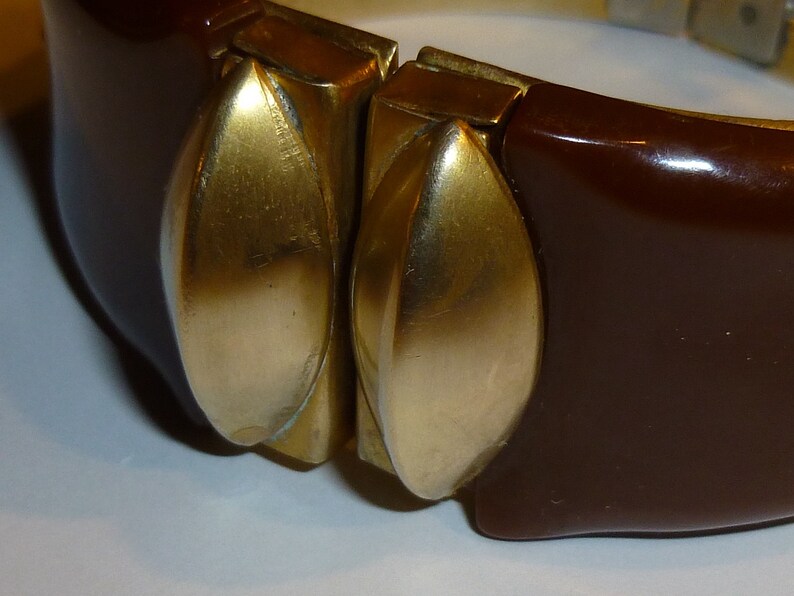 May include: A brown plastic bangle bracelet with two gold-toned metal accents. The accents are shaped like leaves and are attached to the bracelet with a hinge.