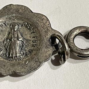 May include: A silver-toned metal charm featuring a scalloped edge and a central embossed figure of a person with a halo. The text O HOLY MARY PRAY FOR US 1830 is visible. A circular spring ring clasp is attached to the side, stamped with STERLING.