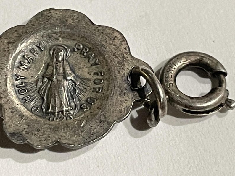 May include: A silver-toned metal charm featuring a scalloped edge and a central embossed figure of a person with a halo. The text O HOLY MARY PRAY FOR US 1830 is visible. A circular spring ring clasp is attached to the side, stamped with STERLING.