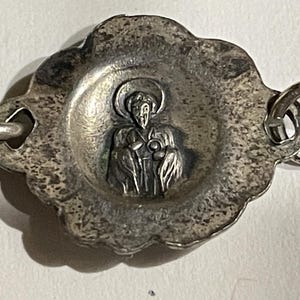 May include: A silver-toned metal charm featuring a central embossed figure of a haloed person in robes. The charm has a scalloped, textured border and is attached to a metal link chain on both sides, displaying a weathered, antique-style finish.