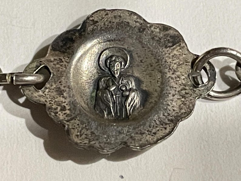 May include: A silver-toned metal charm featuring a central embossed figure of a haloed person in robes. The charm has a scalloped, textured border and is attached to a metal link chain on both sides, displaying a weathered, antique-style finish.