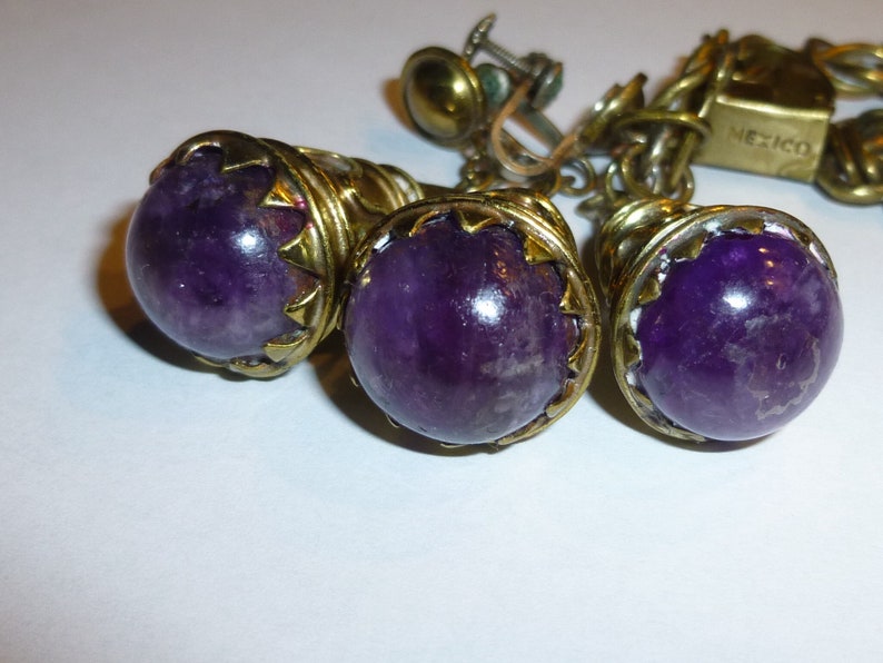 May include: A set of three amethyst earrings with gold-tone metal settings. The earrings are marked "Mexico".