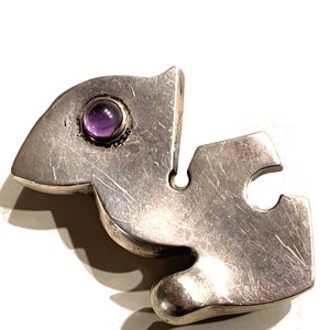 May include: A silver abstract pendant with a purple gemstone set in the center.