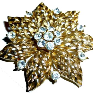 May include: A gold-toned brooch in the shape of a flower with a woven design and clear rhinestones.