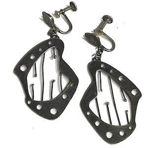 May include: A pair of silver-toned clip-on earrings. Each earring features a geometric, openwork design with a series of small holes around the edge and vertical lines within. The earrings have a screw-back mechanism.