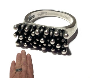 May include: A silver ring with a textured, raised design. The ring has a band that is smooth and the top of the ring is a rectangular shape with a textured design of small, round bumps.