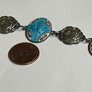 May include: A silver-toned link bracelet featuring alternating circular floral embossed charms and oval turquoise enamel plaques with small pink floral accents, shown next to a copper-coloured penny for scale to indicate size.