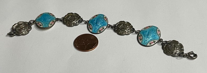 May include: A silver-toned link bracelet featuring alternating circular floral embossed charms and oval turquoise enamel plaques with small pink floral accents, shown next to a copper-coloured penny for scale to indicate size.