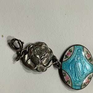 May include: A silver-toned charm bracelet component featuring a three-dimensional rose charm connected to an oval pendant. The pendant displays a blue enameled figure of the Virgin Mary surrounded by white enamel with small pink floral accents.